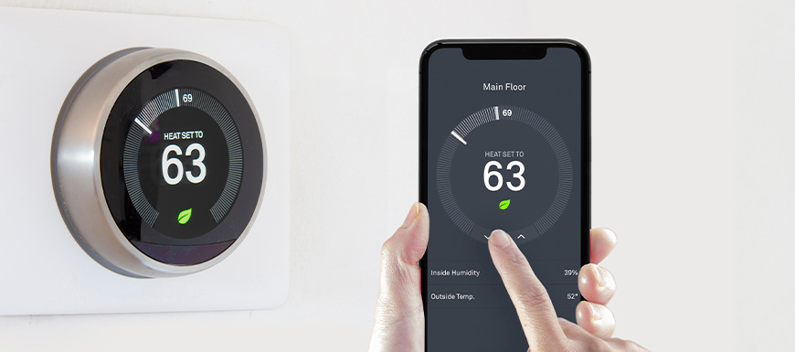 Smart thermostat on wall beside smart thermostat app on phone.